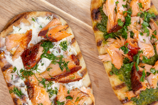 Two flatbreads topped with cream cheese, smoked salmon, and fresh herbs on a wooden cutting board.